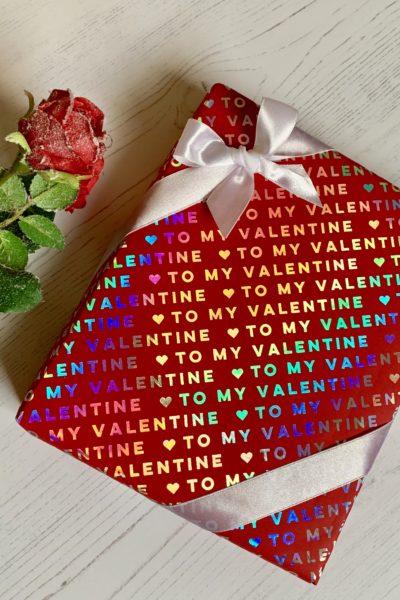 Red giftwrap with 'to my valentine' printed on it in silver foil. A white corner bow and a rose on the side